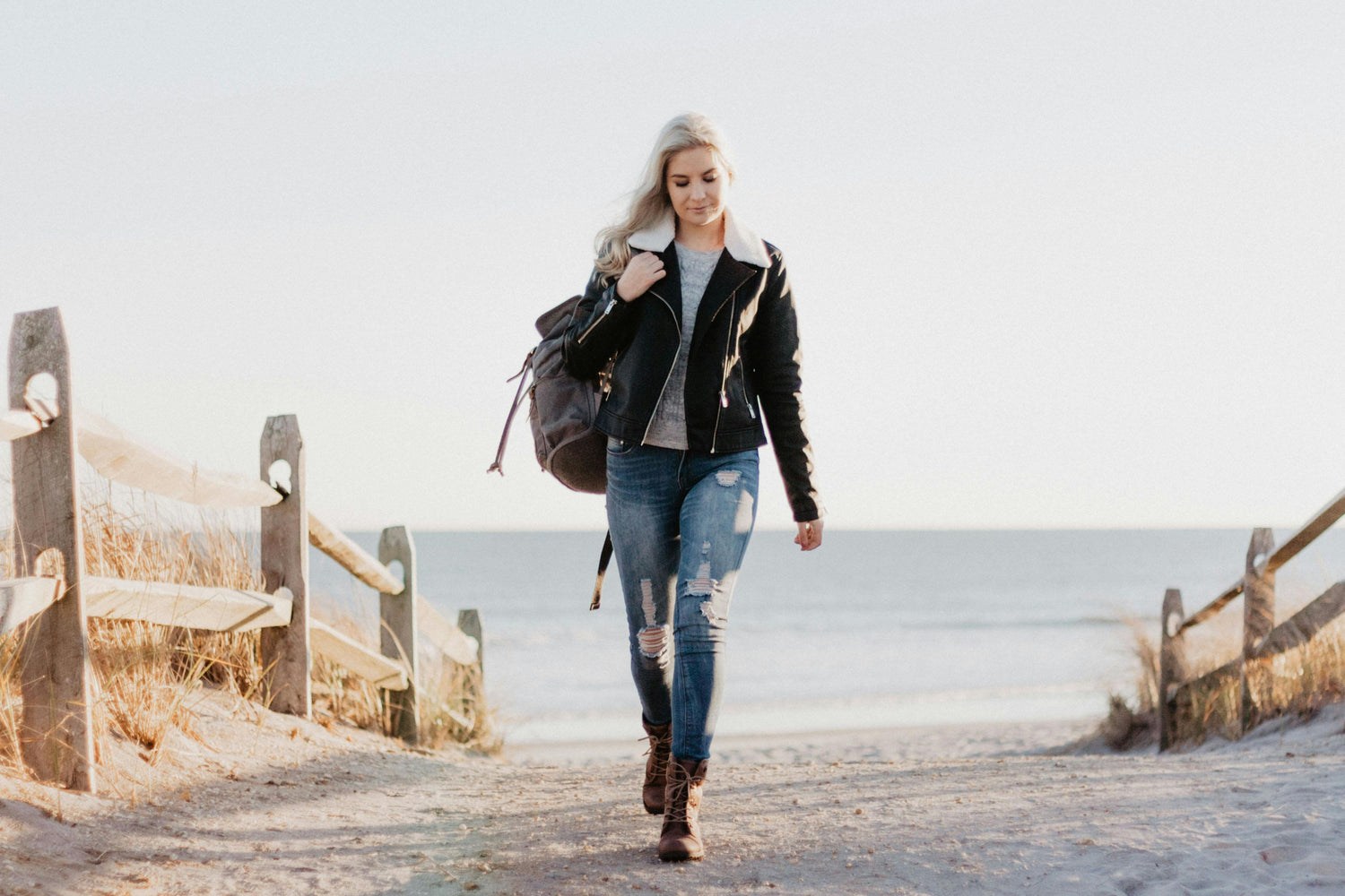 Woman in a leather coat and distressed jeans carrying a backpack over her shoulder walking down a path away from the ocean